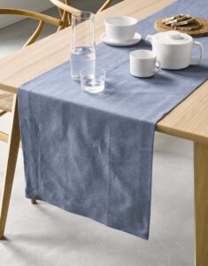 Op een lichte houten tafel ligt de Marc O'Polo Akalla tafelloper in de kleur Smoke Blue, met een karaf, een waterglas, een theepot en gebakjes.