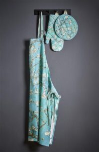 Het Van Gogh Blossom-schort in blauw met witte bloemenprint hangt samen met de bijpassende ovenwant en pannenlap aan een zwart wandrek voor een donkergrijze muur.