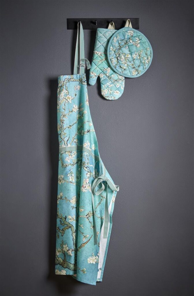 Het Van Gogh Blossom-schort in blauw met witte bloemenprint hangt samen met de bijpassende ovenwant en pannenlap aan een zwart wandrek voor een donkergrijze muur.