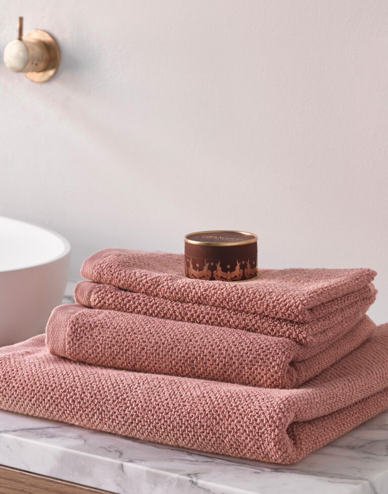 Op een marmeren aanrecht liggen drie opgevouwen Essenza Connect Organic Uni Handdoek Rose met daarbovenop een bruin rond doosje.