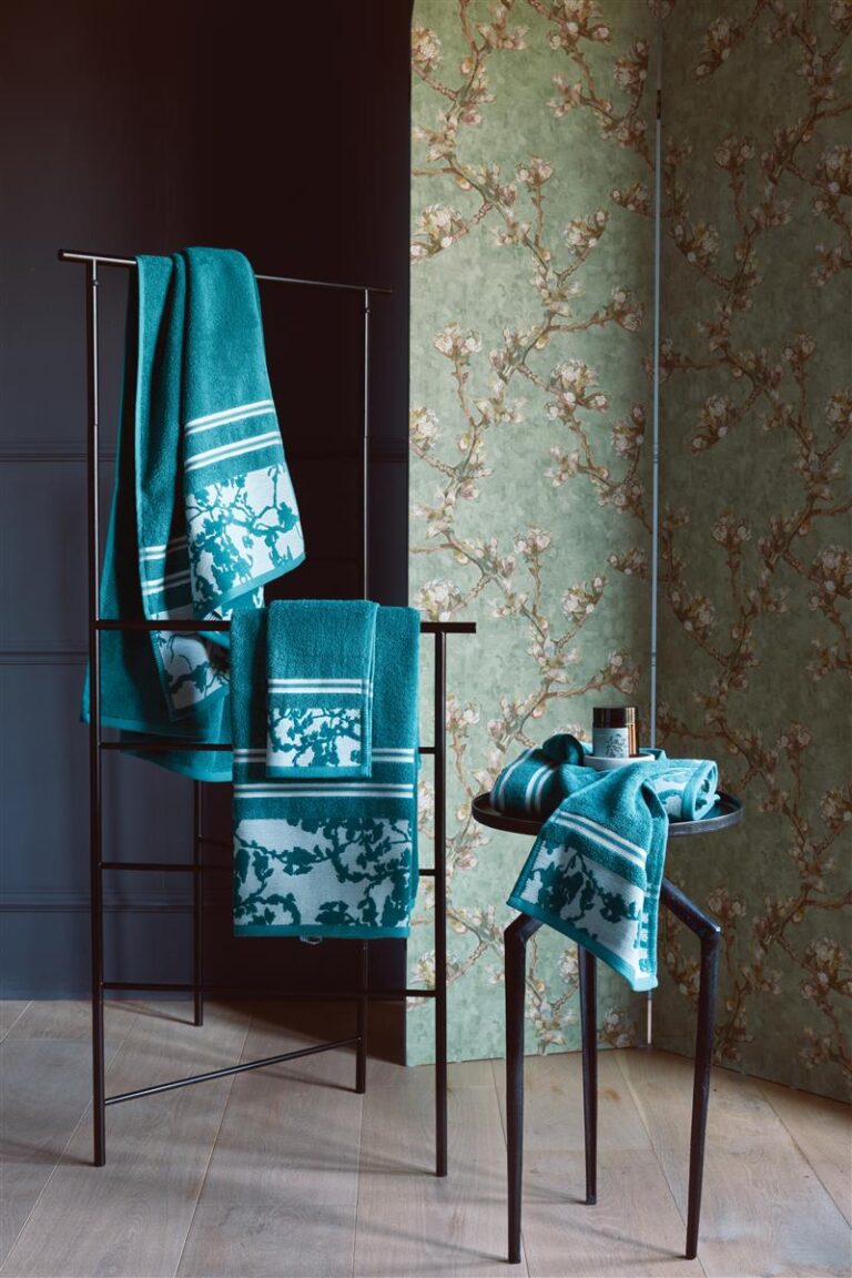 De Van Gogh Fleurir-washandjes in blauw, een set van drie, liggen stijlvol op een zwart handdoekenrek en een tafeltje tegen een bloemenbehang met een donkere muur.