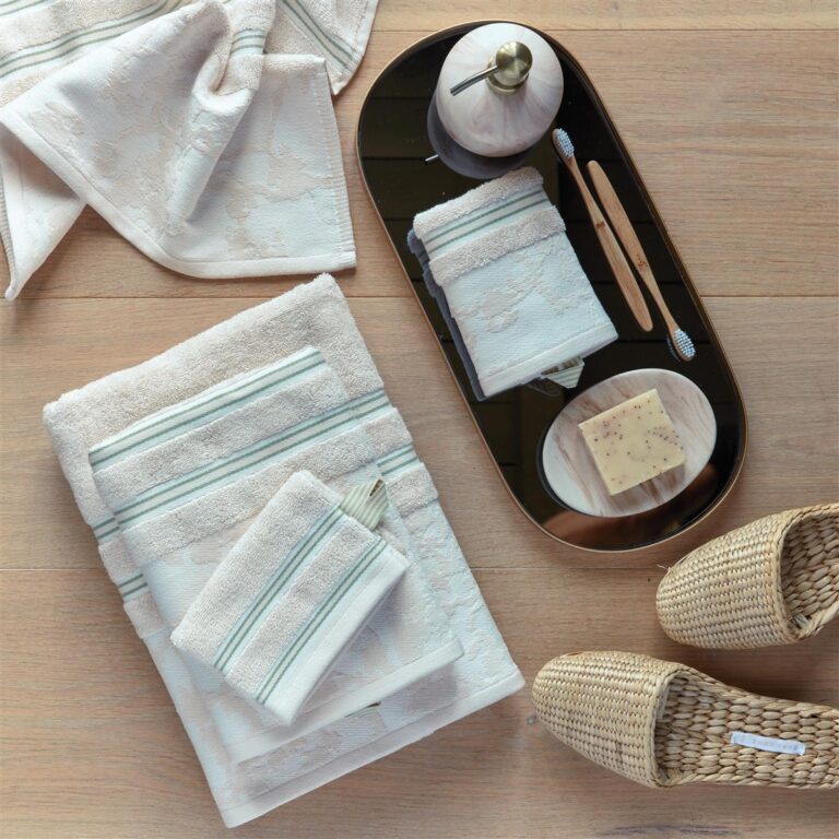 De Van Gogh Fleurir-washandset in gebroken wit bestaat uit drie zachte beige washandjes met groene strepen, een stuk zeep op een schaaltje, een zeepdispenser en gevlochten slippers, die netjes bij elkaar op een lichte houten vloer liggen.