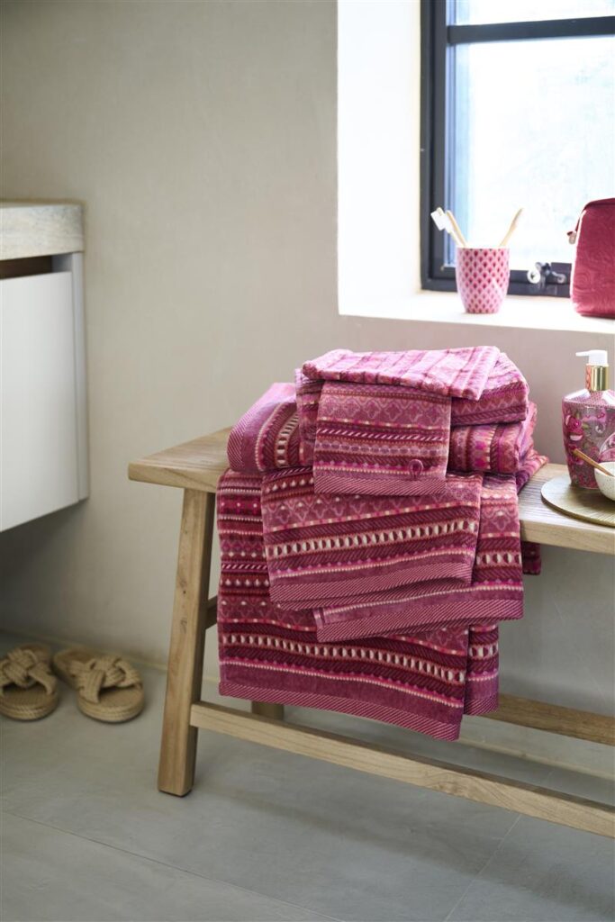 De Pip Studio Geometric gastendoekenset in donkerroze, bestaande uit drie stuks, ligt opgevouwen op een houten bankje bij het raam, naast slippers en een toilettas.