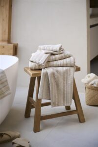 Op een houten kruk liggen gestreepte handdoeken en de Pip Studio Geometric Print Washand Zand-set, met daarnaast slippers en een mandje.