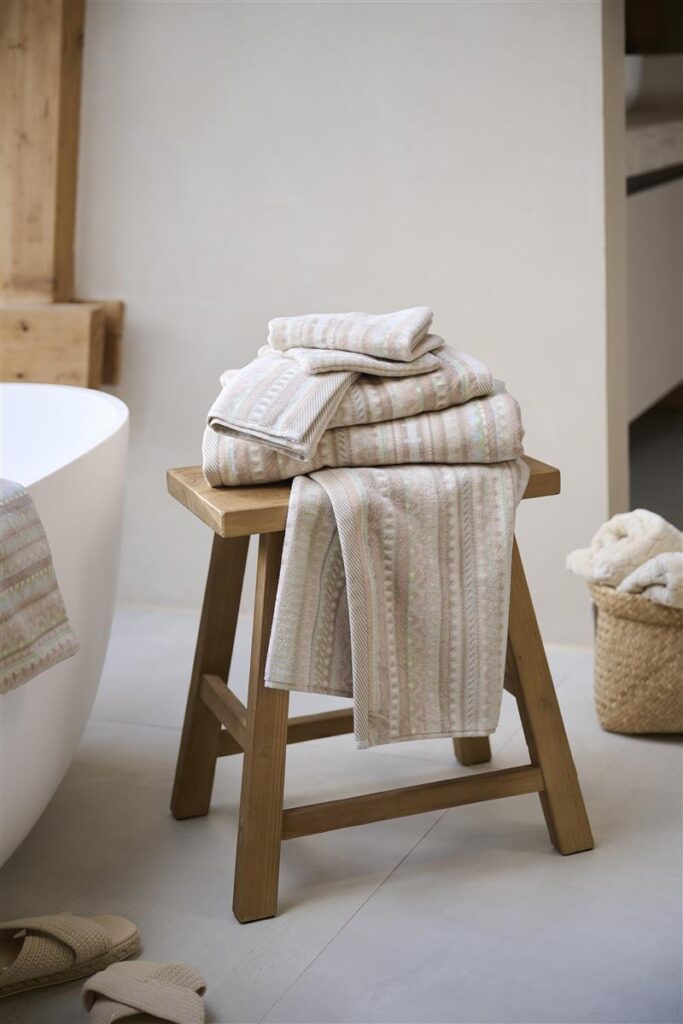 Op een houten kruk liggen gestreepte handdoeken en de Pip Studio Geometric Print Washand Zand-set, met daarnaast slippers en een mandje.