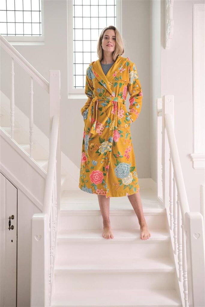 Een persoon staat op blote voeten op witte traptreden en draagt de Pip Studio Good Evening-badjas in geel, maat XL, een gele badjas met bloemenprint, capuchon en zakken.