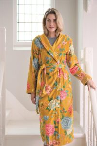 Een vrouw draagt de Pip Studio Good Evening-badjas in geel (maat XL) met grote blauwe, roze en groene bloemen voor een raam met vakken.
