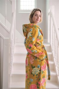 Een vrouw draagt de Pip Studio Good Evening-badjas in geel, maat XL, en kijkt over haar schouder naar de camera terwijl het zonlicht naar binnen schijnt.