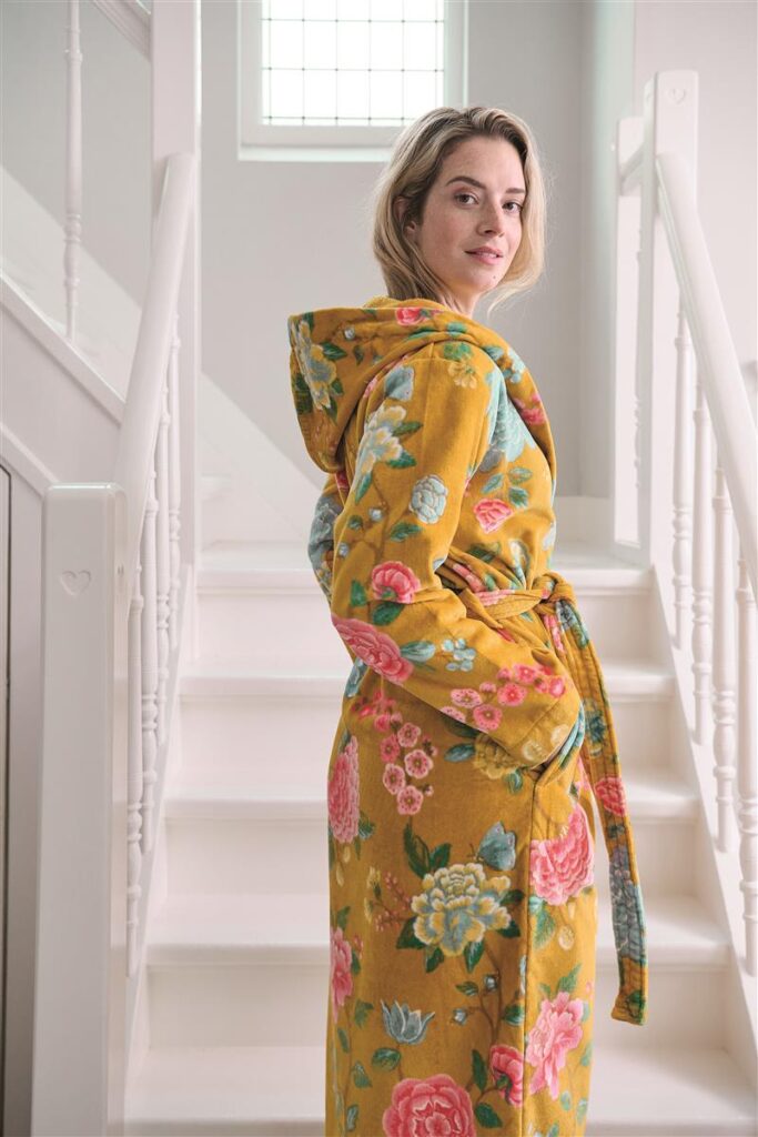 Een vrouw draagt de Pip Studio Good Evening-badjas in geel, maat XL, en kijkt over haar schouder naar de camera terwijl het zonlicht naar binnen schijnt.