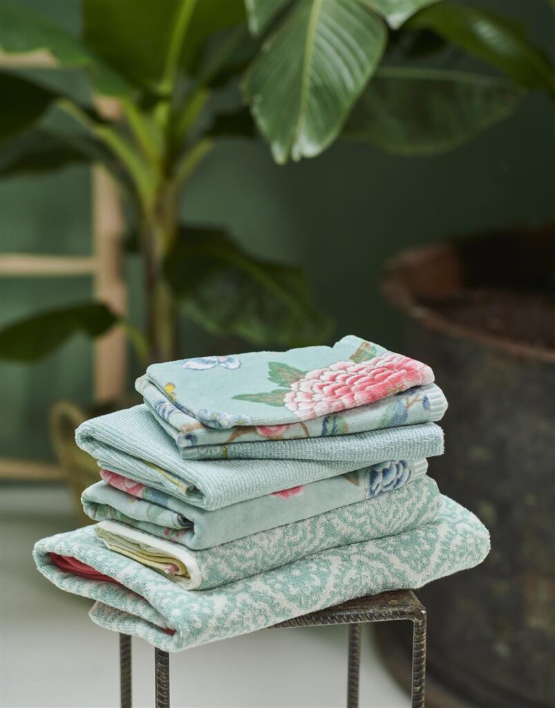 Op een metalen kruk ligt een stapel opgevouwen handdoeken met patronen, zoals de Pip Studio Good Evening Washand Blauw-set, met op de achtergrond zacht vervaagde groene planten.