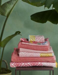 Op een klein tafeltje ligt de Pip Studio Good Evening-set van drie gastendoekjes in koraalrood, omringd door grote groene bladeren.