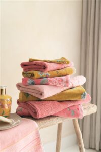 Op een houten bank tegen een beige muur ligt een stapel Pip Studio Good Evening-gastendoekjes in koraalrood met bloemenmotief, samen met een schoonmaakdoekje en een extra doekje.