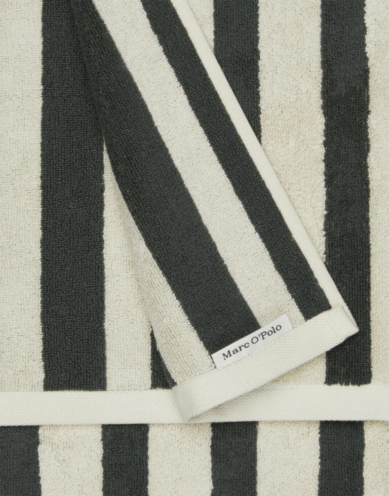 Een close-up van de Marc O'Polo Heritage gastendoek in antraciet toont een zacht, netjes gevouwen zwart-wit gestreept handdoekje met een subtiel opgenaaid Marc O'Polo-label in zwarte letters.