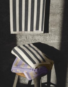 De Marc O'Polo Heritage-servet in lila met opvallende verticale strepen in lila en beige hangt over een kruk bij een betonnen muur.