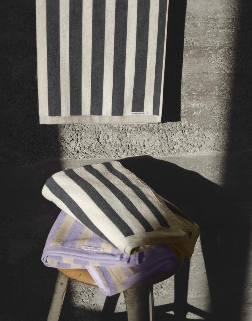 De Marc O'Polo Heritage-servet in lila met opvallende verticale strepen in lila en beige hangt over een kruk bij een betonnen muur.
