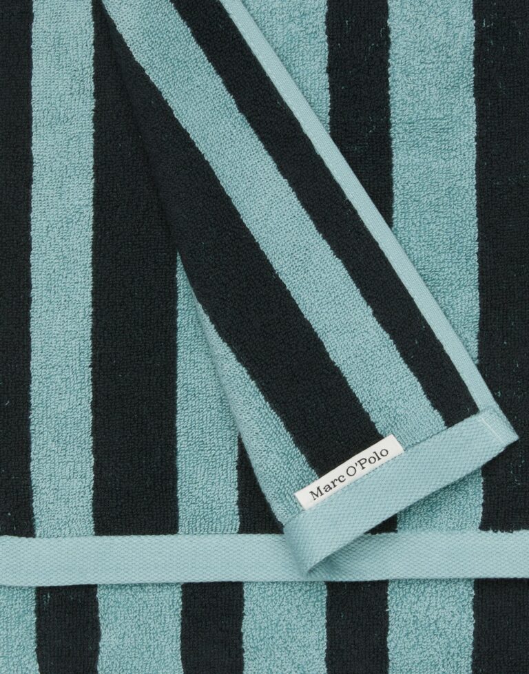 Deze close-up toont de Marc O'Polo Heritage-handdoek in de kleur Aquamarine, met brede zwarte en blauwgroene strepen en een subtiel merklabel aan de rand.