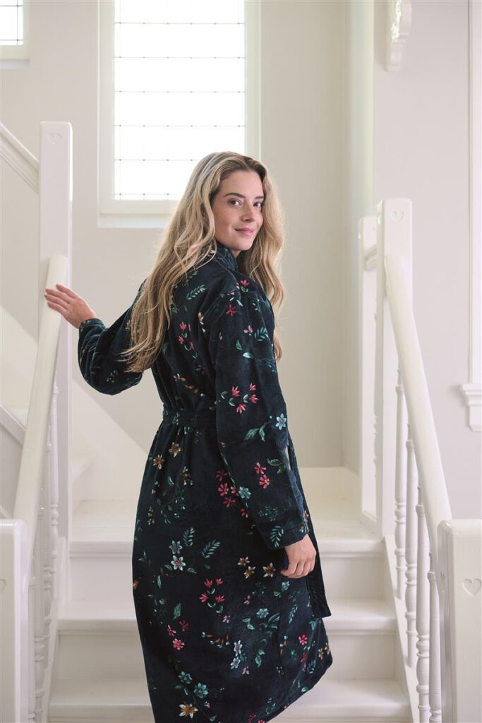 Een vrouw met lang blond haar glimlacht in het zonlicht en draagt de elegante Pip Studio Les Fleurs-badjas in donkerblauw, maat XS.