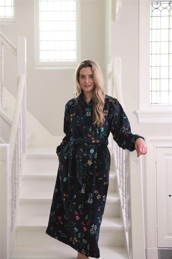 Een vrouw met lang blond haar glimlacht zacht terwijl ze de donkerblauwe Pip Studio Les Fleurs-badjas (XS) draagt.
