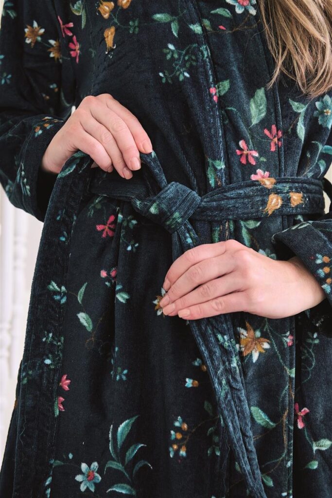 Een persoon draagt de Pip Studio Les Fleurs-badjas in donkerblauw, maat XS, en knoopt de ceintuur om zijn middel, waarbij het elegante bloemenpatroon van de badjas duidelijk zichtbaar is.