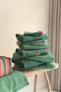 Een set Pip Studio Les Fleurs Gastendoek Groen-handdoeken met bloemenprint ligt op een houten bankje naast een gestreepte tas en een gordijn in een zacht verlichte kamer.