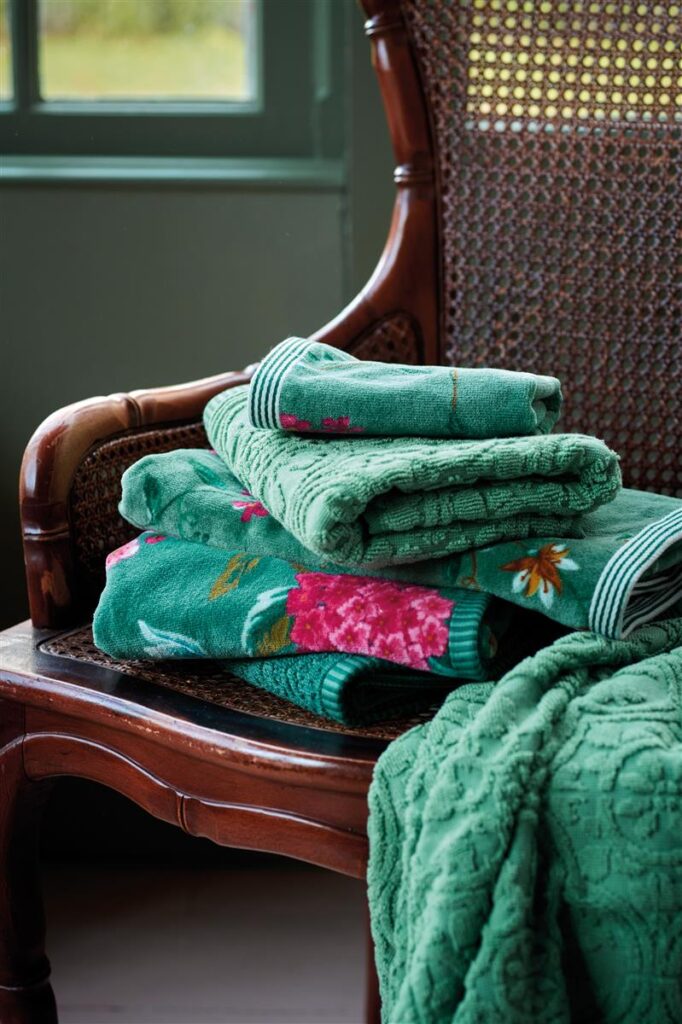 Groene handdoeken met bloemmotieven uit de Pip Studio Les Fleurs Gastendoek Groen-set liggen op een houten stoel met gevlochten rugleuning, terwijl een groene plaid over de armleuning hangt.
