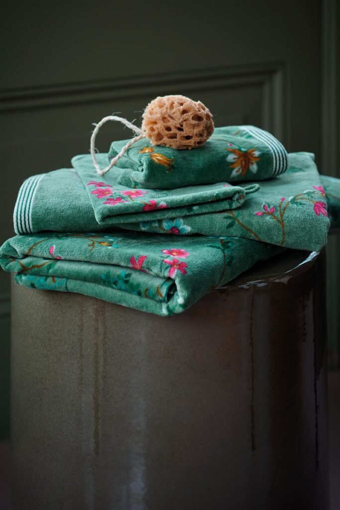 Drie Pip Studio Les Fleurs-handdoeken in groen, met roze bloemen en witte strepen, voorzien van een natuurspons op een glanzend oppervlak tegen een groene achtergrond.