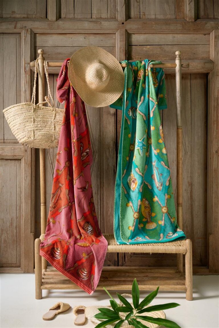 Het Pip Studio Little Sea-strandlaken in groen hangt samen met een ander strandlaken over een houten rek in een zomerse setting met een strohoed, een gevlochten tas, slippers en verse groene bladeren tegen een houten wand.