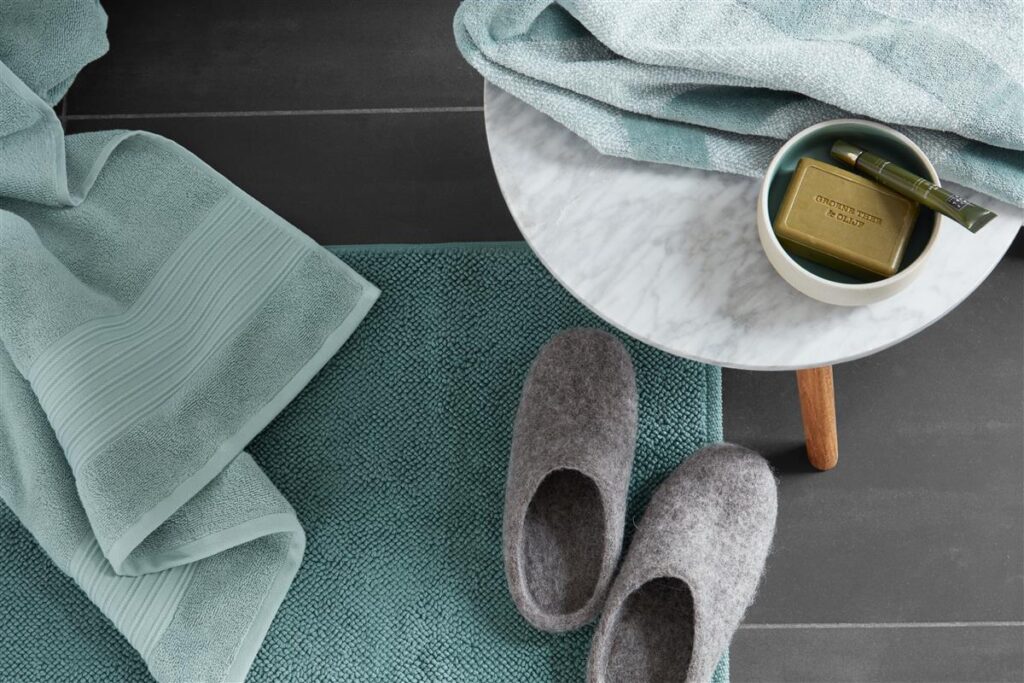 In de stijlvolle badkamer liggen lichtblauwe handdoeken en grijze badslippers, en op een ronde marmeren tafel staat groene zeep in een schaal op donkere tegels, met op de vloer de Beddinghouse Nova badmat Groen voor een verfijnde uitstraling.