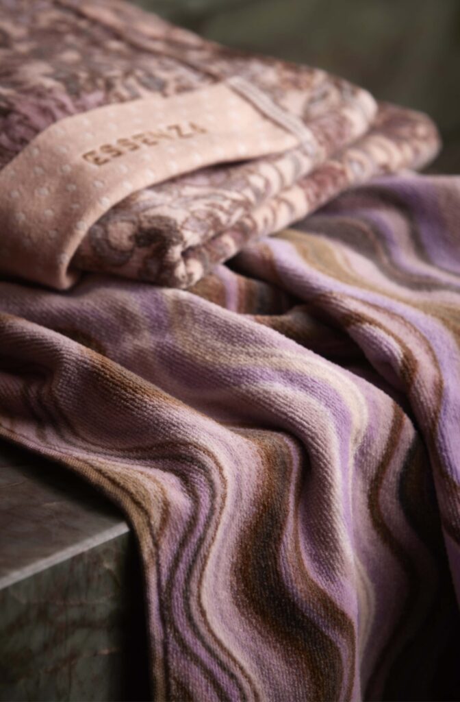 Twee opgevouwen Essenza Ophelia-handdoeken in de kleur Darling Pink liggen op marmer, met zachte patronen in roze, beige, paars en bruin.