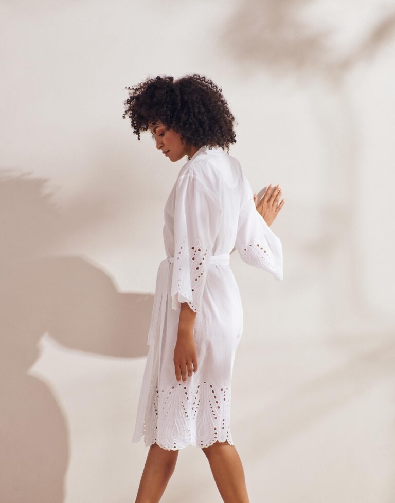Een vrouw met krullend haar staat zijdelings en draagt de Essenza Sarai Tilia Kimono in Pure White met kanten details aan de mouwen en de zoom.