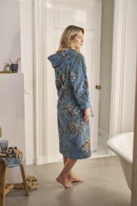 Een vrolijke vrouw op blote voeten draagt de blauwe, met bloemen versierde Pip Studio Secret Garden-badjas (blauw, maat XS) in een badkamer met bad en plank.