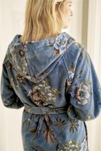 Een vrouw met blond haar is van achteren te zien terwijl ze binnenshuis de blauwe, met bloemen versierde Pip Studio Secret Garden-badjas draagt.