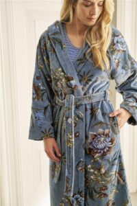 Een vrouw draagt binnenshuis de Pip Studio Secret Garden-badjas in blauw (maat XS) over een gestreept shirt, met een hand in haar zak. De badjas heeft een ceintuur in de taille en is versierd met een elegant bloemenpatroon.
