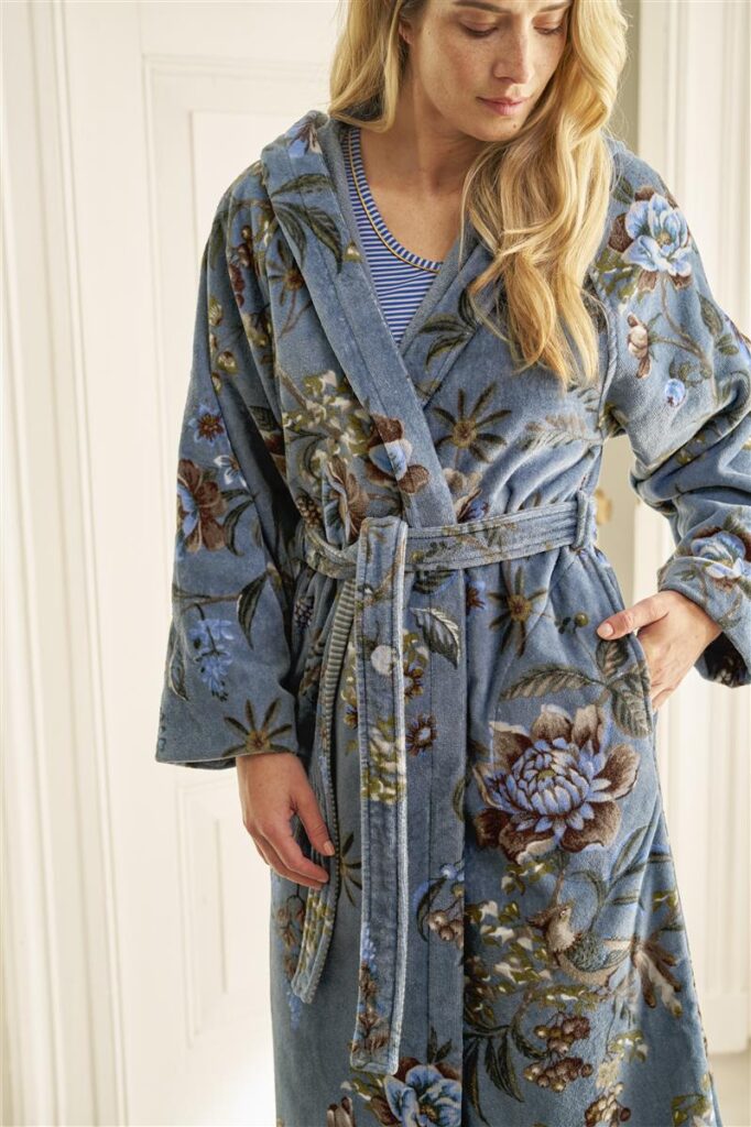 Een vrouw draagt binnenshuis de Pip Studio Secret Garden-badjas in blauw (maat XS) over een gestreept shirt, met een hand in haar zak. De badjas heeft een ceintuur in de taille en is versierd met een elegant bloemenpatroon.