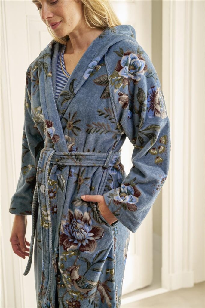 Een vrouw draagt de Pip Studio Secret Garden-badjas Blauw XS, een blauwe badjas met bloemenprint, capuchon en ceintuur, en glimlacht subtiel terwijl ze haar hand in haar zak steekt.