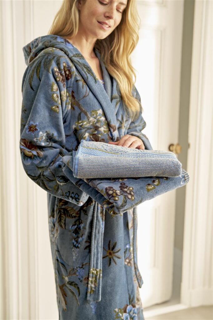 Een glimlachende vrouw met lang blond haar draagt de Pip Studio Secret Garden-badjas in blauw (maat XS) en houdt een opgevouwen blauwe handdoek vast.