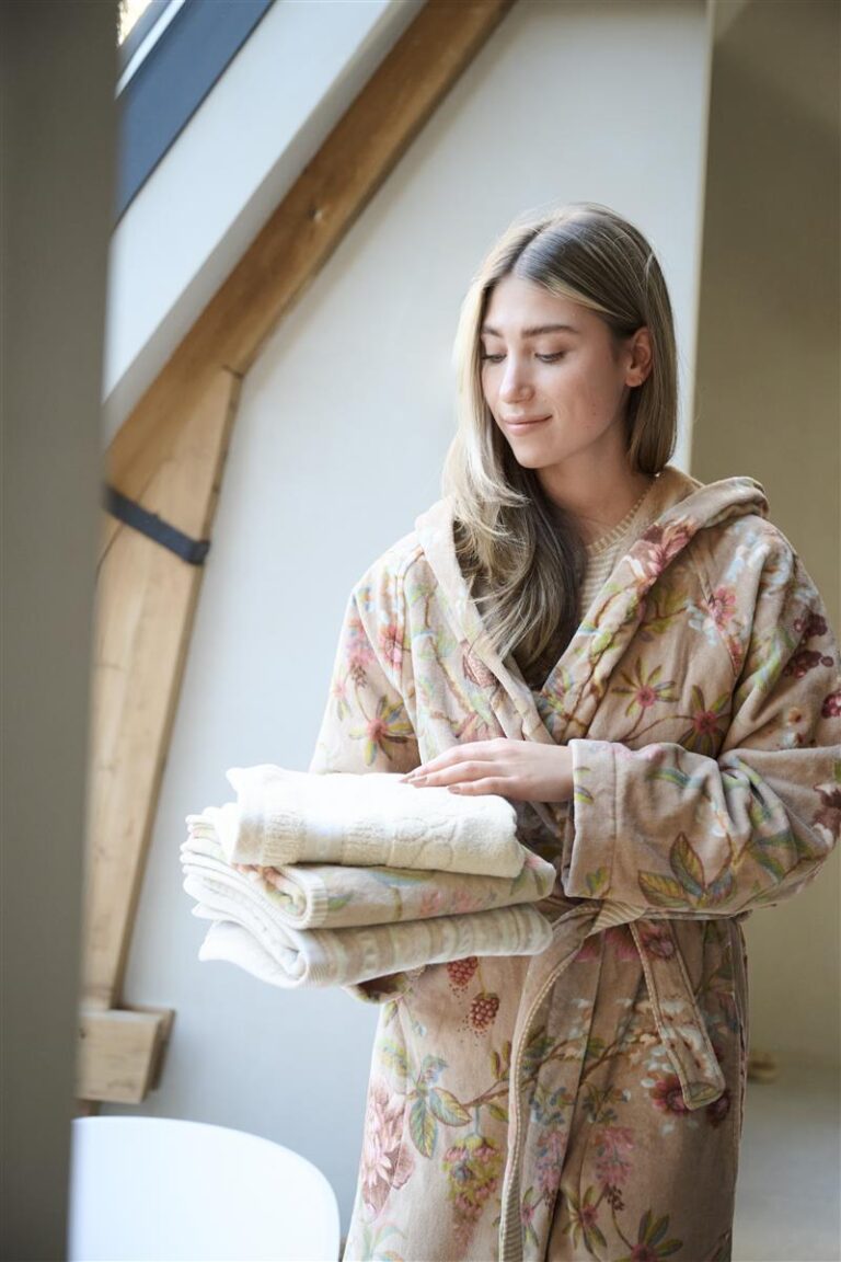 Een vrouw staat binnen bij het raam in een Pip Studio Secret Garden-badjas in de kleur khaki (maat XS) en glimlacht terwijl ze handdoeken vasthoudt.