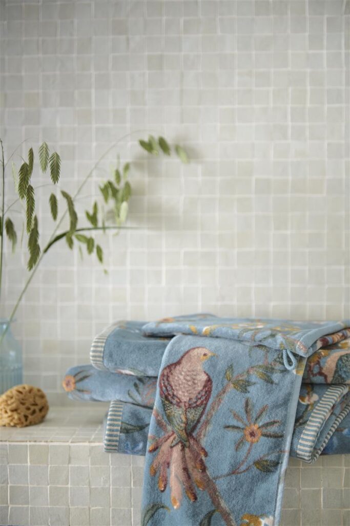De handdoeken uit de Pip Studio Secret Garden Washand Blauw-collectie, met geborduurde vogels en takken, liggen opgestapeld naast een glazen vaas met groene twijgen en een natuurspons, sfeervol gepresenteerd tegen een lichte muur.