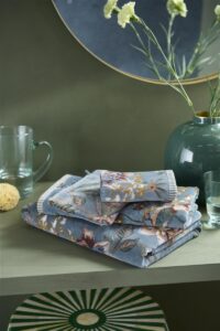 De Pip Studio Secret Garden Washand Blauw-set met bloemenprint staat op een groene plank naast glaswerk, een vaas en gele bloemen.