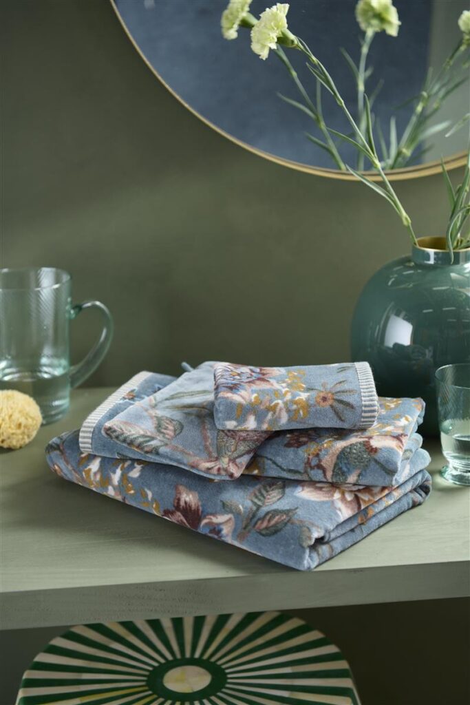De Pip Studio Secret Garden Washand Blauw-set met bloemenprint staat op een groene plank naast glaswerk, een vaas en gele bloemen.