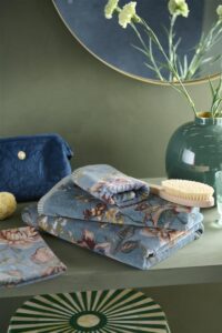 Op een groene plank ligt een stapel blauwe handdoeken met bloemenprint van Pip Studio Secret Garden Washand Blauw, naast verzorgingsproducten.