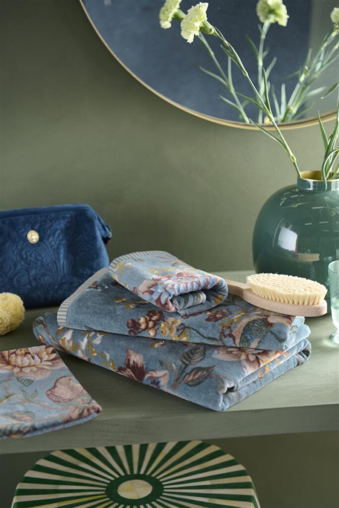Op een groene plank ligt een stapel blauwe handdoeken met bloemenprint van Pip Studio Secret Garden Washand Blauw, naast verzorgingsproducten.