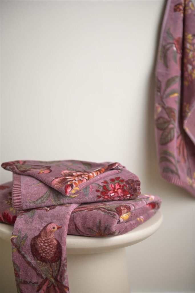 Op een witte kruk ligt een stapel mauvekleurige Pip Studio Secret Garden Washand Lichtpaars-washandjes met bloemen en vogels, met een bijpassende handdoek die ernaast hangt.