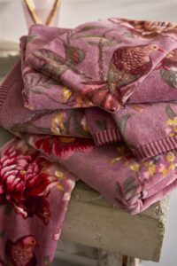 Een stapel zachte, roze handdoeken met bloemen- en vogelmotief ligt op een stenen ondergrond, met een bijpassende washand van Pip Studio nonchalant over de rand gedrapeerd, zodat het verfijnde ontwerp goed tot zijn recht komt.