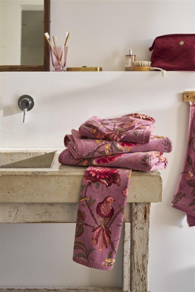 Op een rustieke wastafel ligt een stapel Pip Studio Secret Garden-washandjes in lichtpaars met roze bloemen en vogels, omringd door toiletartikelen en een bordeauxrode tas.