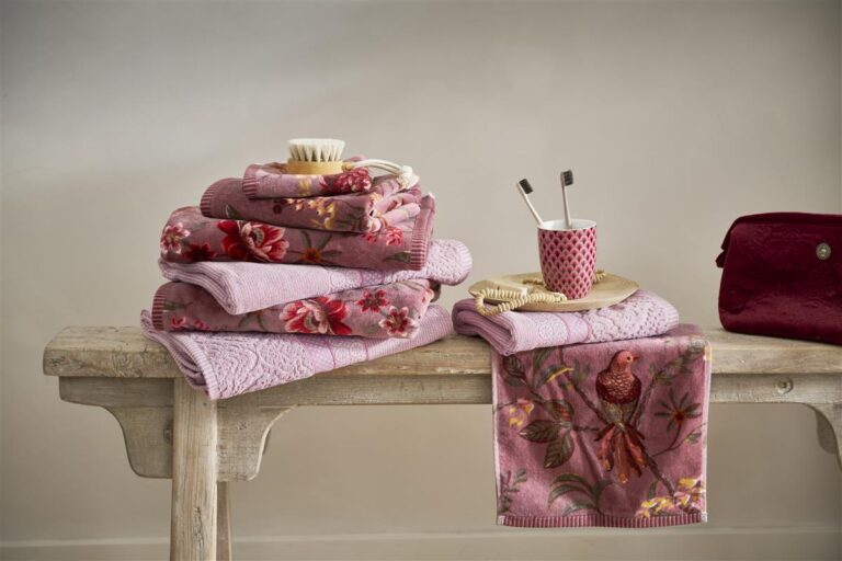 De Pip Studio Secret Garden-washand in lichtpaars met bloemen en vogels ligt op een houten bankje naast borstels, een beker en een toilettas.