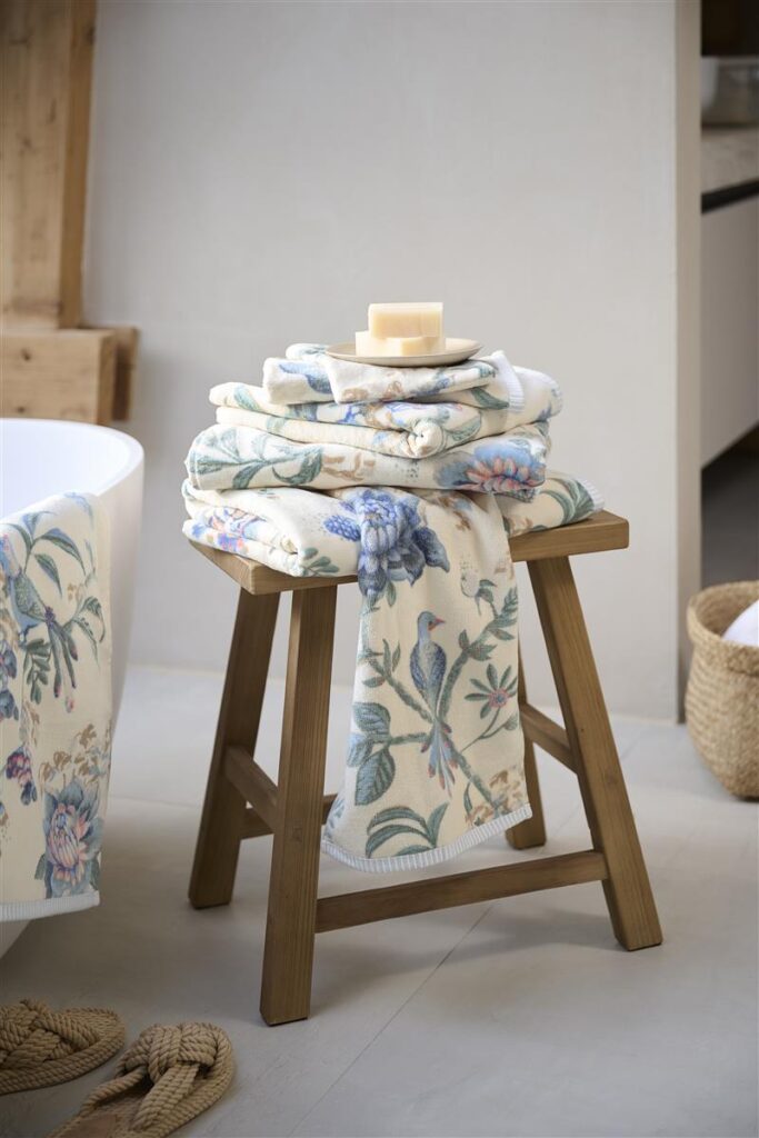 Drie blauw-witte handdoeken van Pip Studio uit de Secret Garden-collectie liggen op een houten krukje met een stuk zeep erop; er hangt ook een bijpassende handdoek bij.
