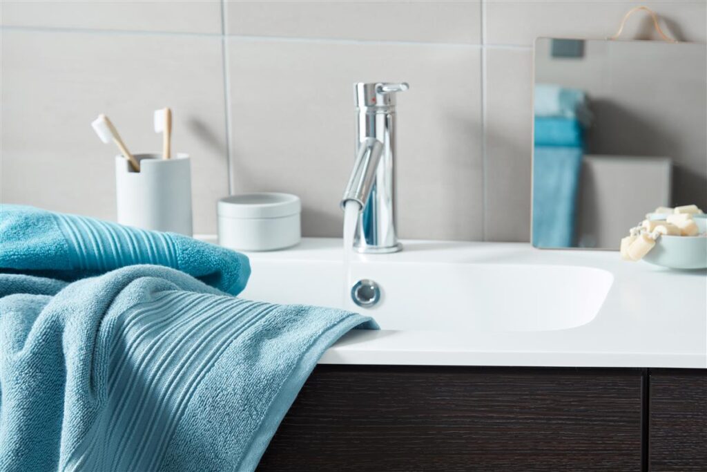 Een moderne badkamer met een chromen kraan waar water uit stroomt, Beddinghouse Sheer Gastendoek Blauw handdoeken op het aanrecht, een tandenborstelhouder en een kleine spiegel aan de betegelde muur.