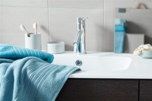 Een moderne badkamer met een chromen kraan waar water uit stroomt, Beddinghouse Sheer Gastendoek Blauw handdoeken op het aanrecht, een tandenborstelhouder en een kleine spiegel aan de betegelde muur.