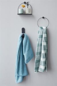 Twee Beddinghouse Sheer Gastendoek Blauw-handdoeken hangen aan een lichtgrijze muur onder een zwarte plank met accessoires.